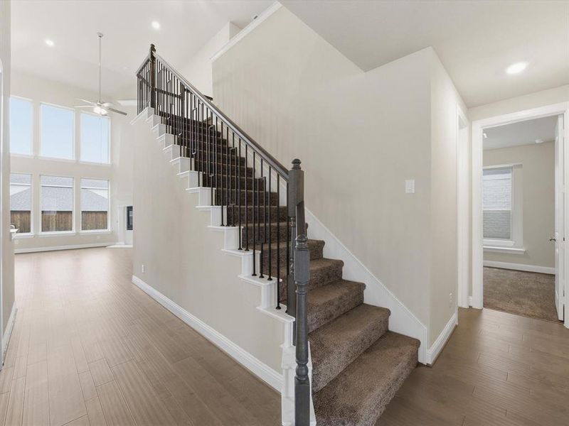 Two-story foyer featuring wood-finish flooring and a carpeted staircase with dark wood banister and metal spindles Two-story foyer featuring wood-finish flooring and a carpeted staircase with dark wood banister and metal spindles