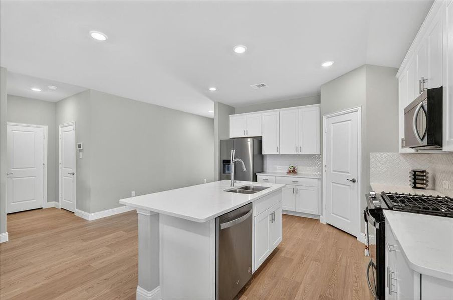 Kitchen featuring a central island with an undermount sink, stainless steel appliances, white cabinetry, light countertops, and wood-finish flooring Kitchen featuring a central island with an undermount sink, stainless steel appliances, white cabinetry, light countertops, and wood-finish flooring