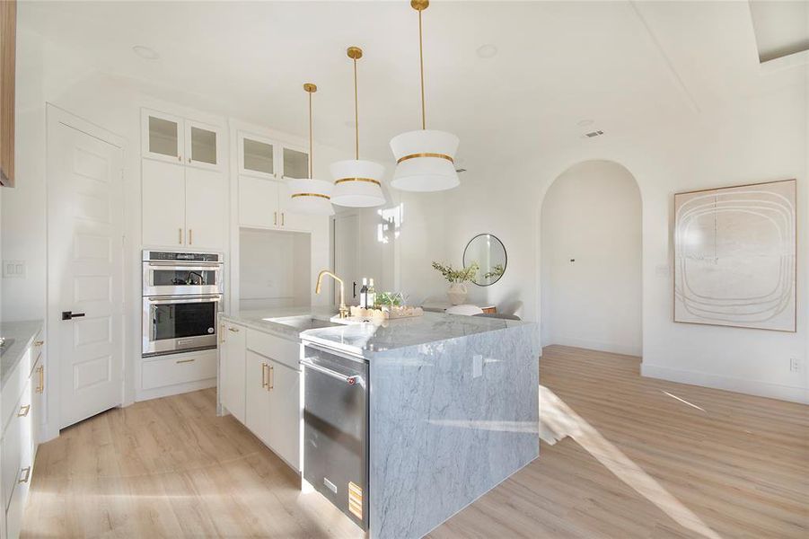 Kitchen featuring glass insert cabinets, white cabinetry, hanging light fixtures, light stone countertops, and an island with sink Kitchen featuring glass insert cabinets, white cabinetry, hanging light fixtures, light stone countertops, and an island with sink