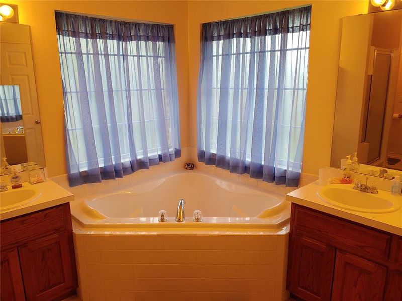 Corner soaking tub with dual window exposures, flanked by two separate vanities featuring wood cabinetry and integrated sinks