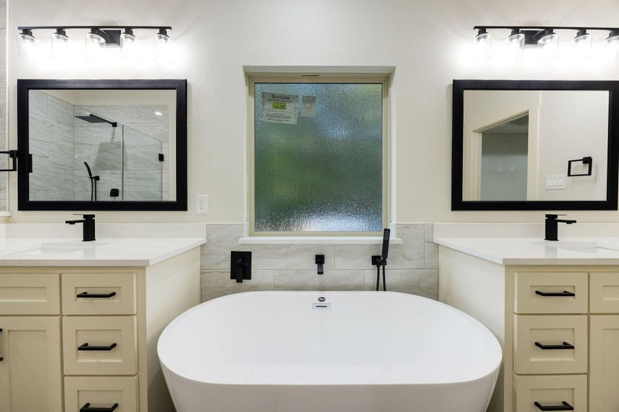 The primary  bathroom features a modern, elegant design with a central freestanding bathtub, dual vanities with black fixtures, and framed mirrors. It includes a frosted window for privacy, offering a serene and stylish space.