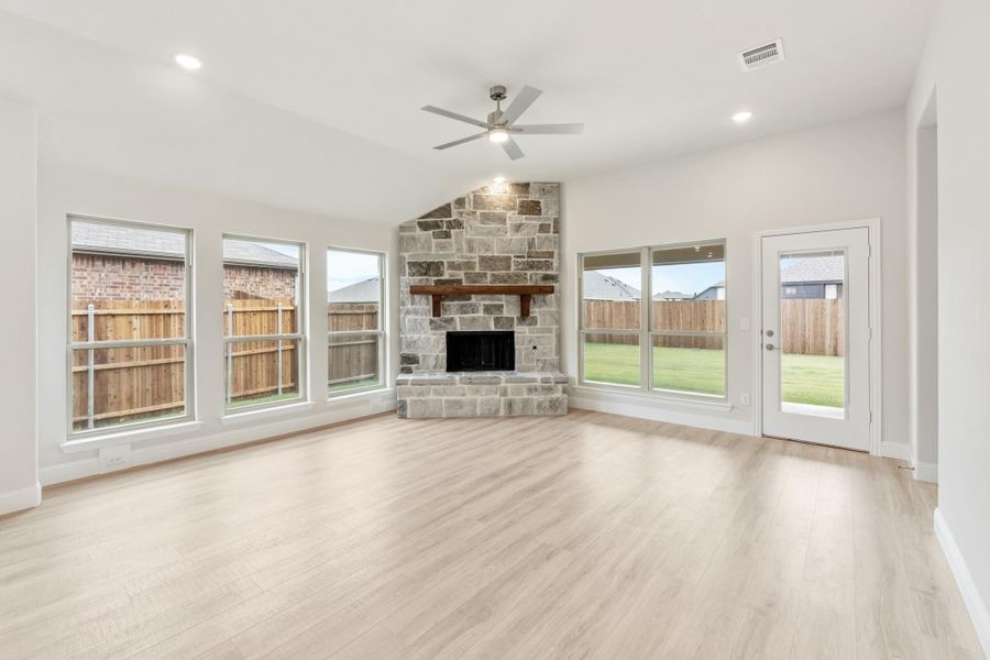 Open living room with stone fireplace, light wood floors, ceiling fan, and large windows
