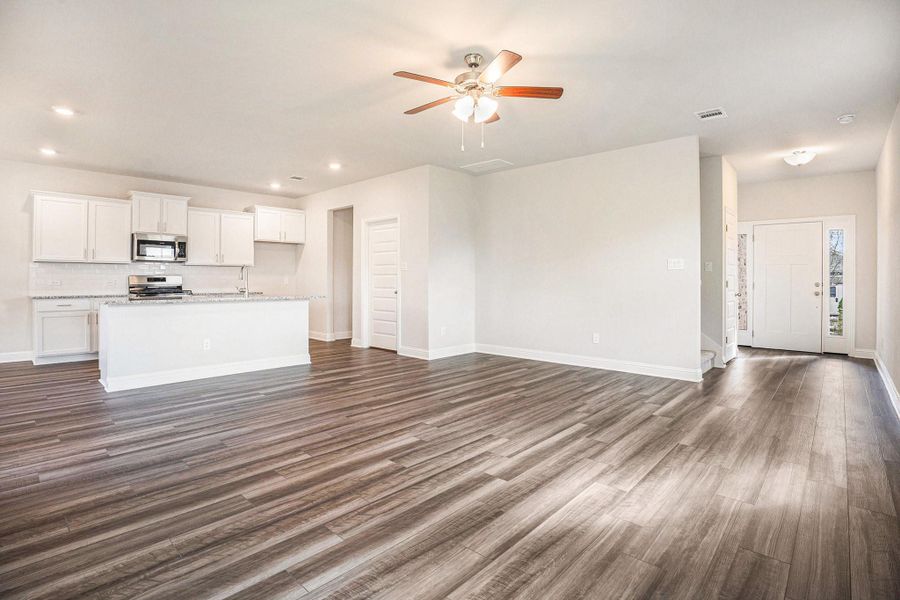 This photo showcases a spacious open-concept living area with luxury vinyl plank flooring and neutral walls. The kitchen features white cabinetry, a large island, and stainless steel appliances. A ceiling fan and recessed lighting enhance the bright and airy feel, while the entryway offers a welcoming touch.