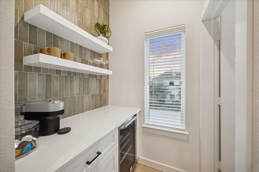 Charming coffee or wine nook with quartz countertop, open shelving, and a built-in beverage fridge. Textured tile backsplash and natural light create a cozy yet refined space for daily routines or entertaining prep.