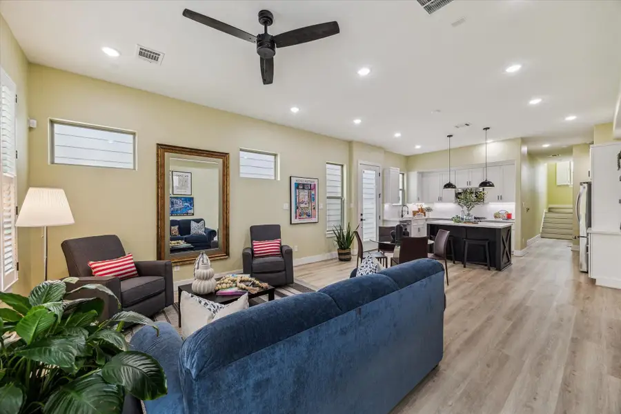 This open-concept living space features modern lighting and a ceiling fan, with a cozy seating area and stylish kitchen. The room is well-lit with natural light from multiple windows, and there's a mix of contemporary and comfortable furnishings.