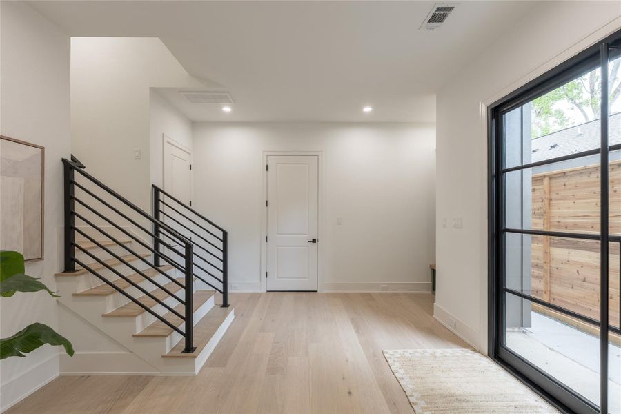 The arrival moment done right; a horizontal steel-balustrade staircase in matte black riding over white oak treads, wide-plank oak flooring underfoot, and a full-height black-framed steel window pulling the cedar courtyard inside. Clean lines, crisp baseboards, and a sightline that lets the architecture do the talking.