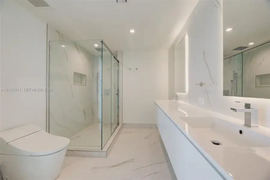 Bathroom featuring a dual-sink vanity with a solid-surface countertop, walk-in glass-enclosed shower with wall-mounted plumbing fixtures, recessed shower niche, and large-format porcelain tile flooring
