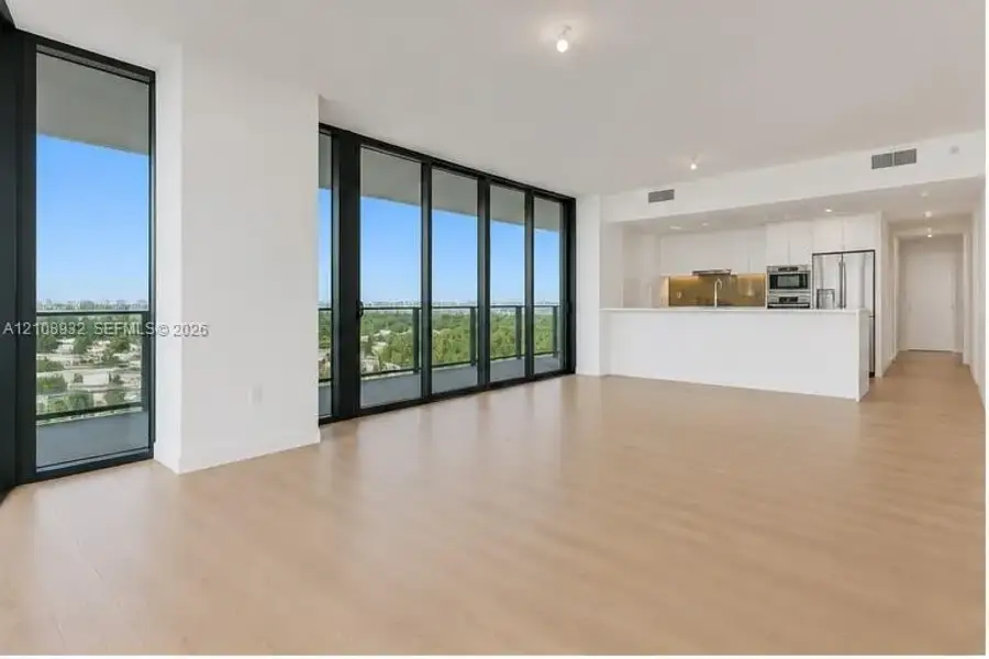 Open-concept living space featuring floor-to-ceiling windows with dark frames, wood-finish flooring, and a kitchen equipped with white flat-panel cabinetry and stainless steel appliances