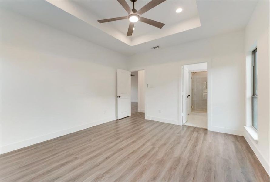 Unfurnished bedroom featuring a raised ceiling, ensuite bathroom, light wood-type flooring, and ceiling fan