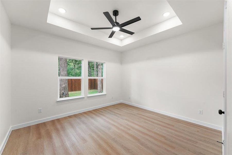 Spacious room featuring a tray ceiling with recessed lighting, a contemporary black ceiling fan, two windows providing natural light, wood-finish flooring, and crisp white trim Spacious room featuring a tray ceiling with recessed lighting, a contemporary black ceiling fan, two windows providing natural light, wood-finish flooring, and crisp white trim