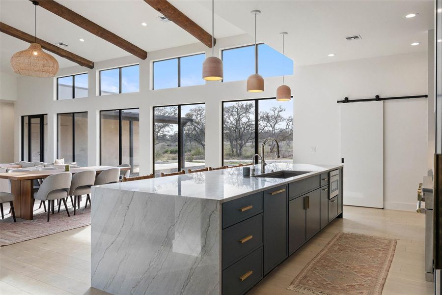 Kitchen featuring a barn door, decorative light fixtures, light stone countertops, a kitchen island with sink, and dark cabinets