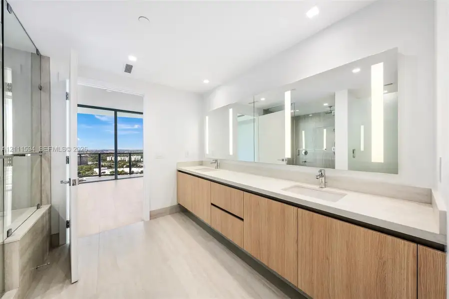 Floating vanity with wood-finish cabinetry, dual under-mount sinks, expansive wall-to-wall integrated mirror with vertical task lighting, and glass-enclosed shower area Floating vanity with wood-finish cabinetry, dual under-mount sinks, expansive wall-to-wall integrated mirror with vertical task lighting, and glass-enclosed shower area