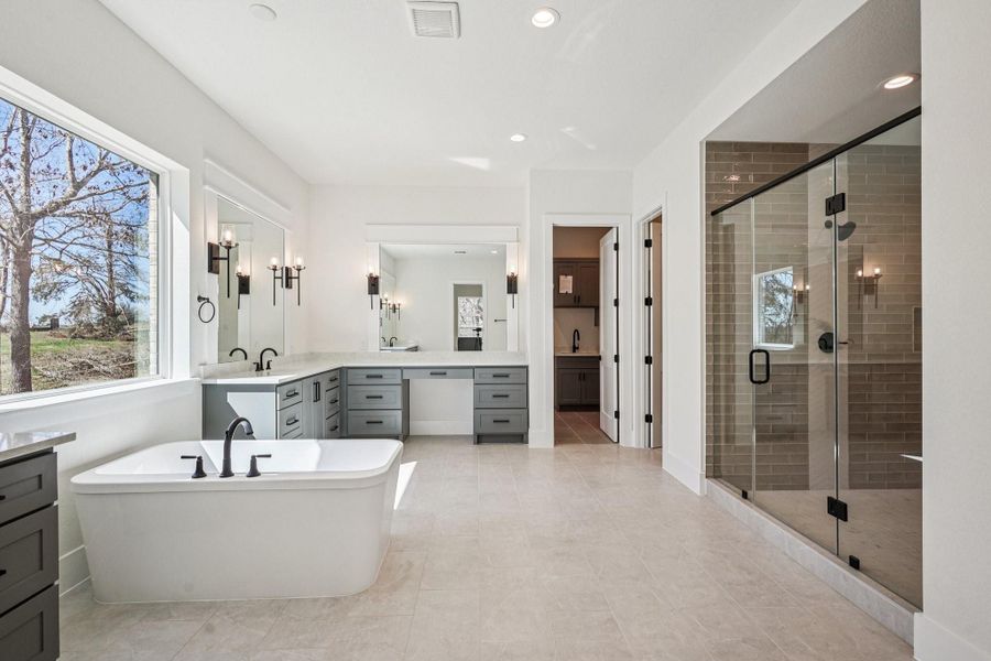 This spacious bathroom features a modern design with a large soaking tub, dual vanities with ample storage, and a glass-enclosed shower. Natural light streams through a generous window, creating a bright and inviting atmosphere.