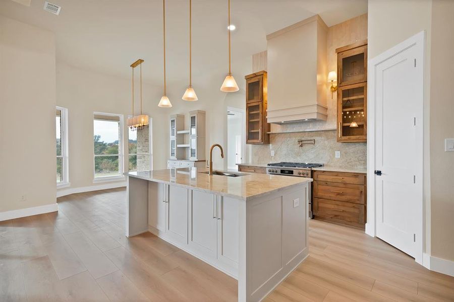 Kitchen featuring glass insert cabinets, hanging light fixtures, light stone countertops, a kitchen island with sink, and tasteful backsplash Kitchen featuring glass insert cabinets, hanging light fixtures, light stone countertops, a kitchen island with sink, and tasteful backsplash