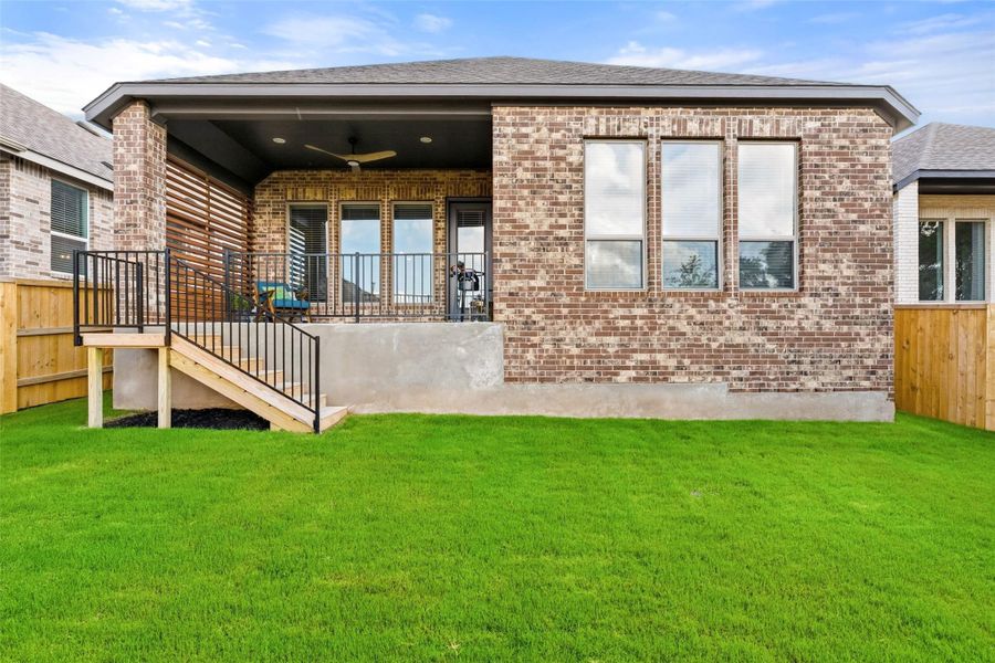 Expansive green lawn with a covered patio featuring a ceiling fan, access stairs with black metal railings, and a brick exterior facade with multiple large windows