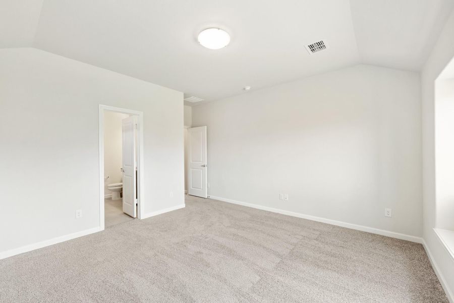 Empty bedroom with vaulted ceiling, carpet flooring, and an open ensuite bathroom door