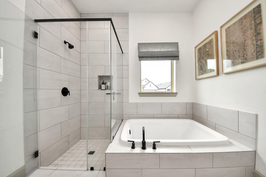 Glass-enclosed shower with sleek black fixtures and a built-in niche. Adjacent is a large, tiled bathtub under a window, allowing natural light to brighten the space. Two framed artworks add a touch of elegance.