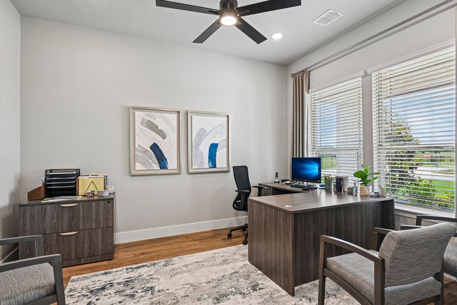 Home office with dark wood desk, ceiling fan, hardwood floors, and large window with blinds