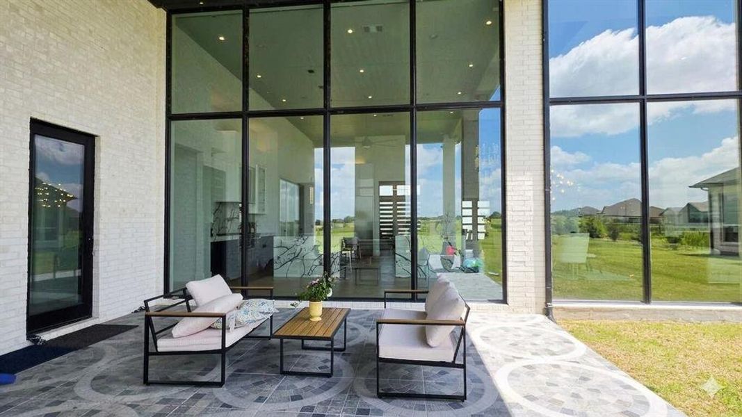Expansive outdoor patio featuring a patterned paver surface, white brick exterior, black-framed floor-to-ceiling windows, a glass-paned door, and a grass lawn