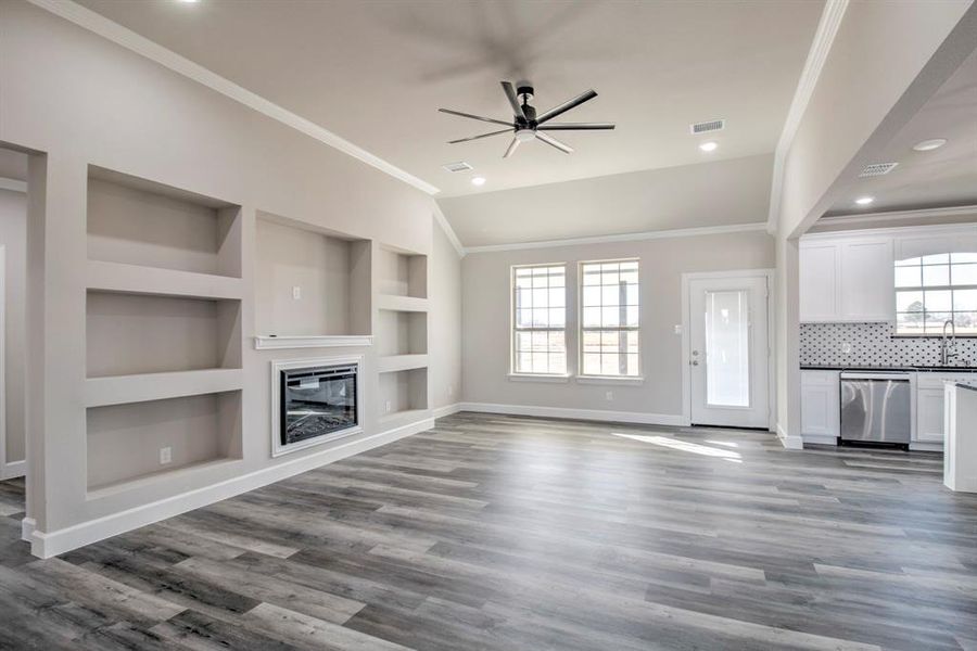 Unfurnished living room featuring built in shelves, ornamental molding, a glass covered fireplace, ceiling fan, and dark wood-style floors