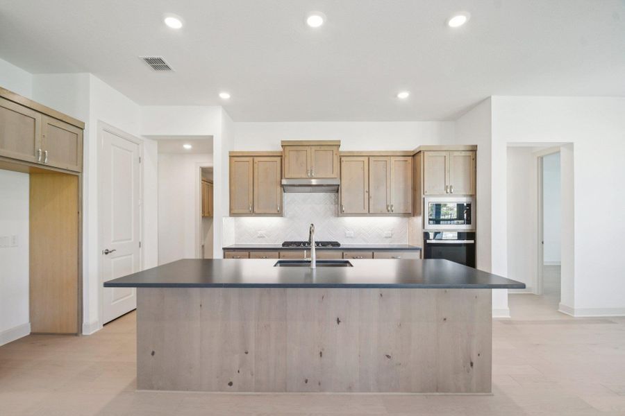 Kitchen with dark countertops, backsplash, a center island with sink, light brown cabinets, and recessed lighting Kitchen with dark countertops, backsplash, a center island with sink, light brown cabinets, and recessed lighting