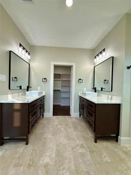 This is a spacious bathroom featuring dual vanities with dark wood cabinets, large mirrors, and modern light fixtures. The neutral-toned tile floor and a walk-in closet add functionality and style. This is a spacious bathroom featuring dual vanities with dark wood cabinets, large mirrors, and modern light fixtures. The neutral-toned tile floor and a walk-in closet add functionality and style.