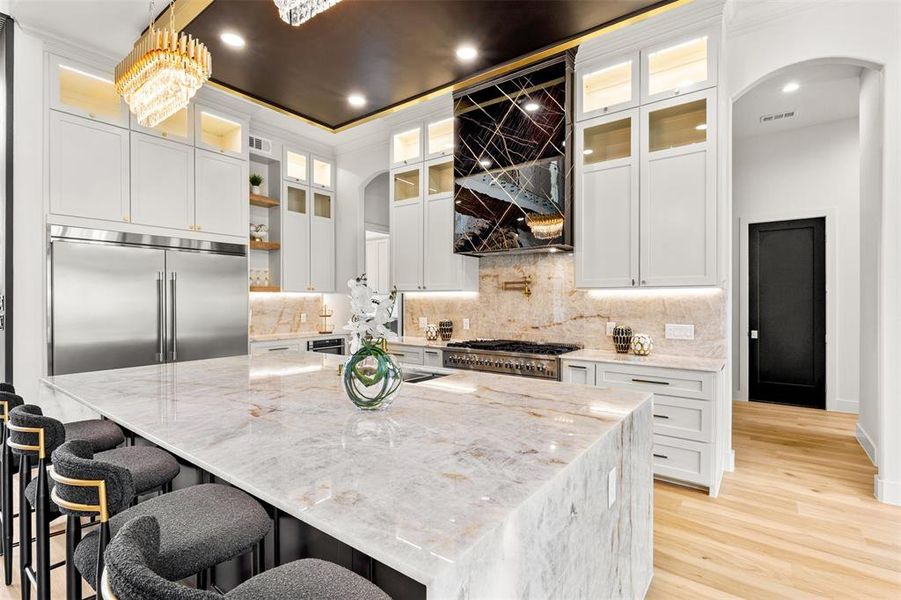 Gourmet kitchen featuring a large stone island with seating, stainless steel built-in refrigerator, professional-grade range, full-height stone backsplash, and extensive white cabinetry with gold-toned hardware