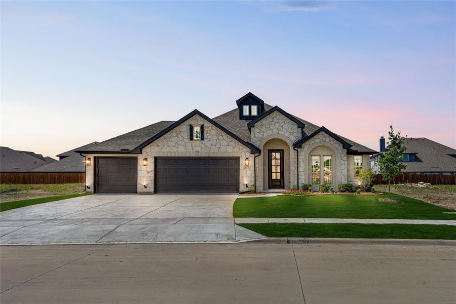 Single-story home with stone exterior, three-car garage, arched entry, and green front lawn at dusk