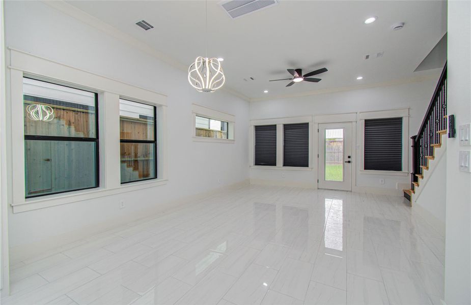 Bright and modern living area with sleek tile flooring, large windows, and a stylish light fixture. Features include a ceiling fan, a door leading to the covered patio and backyard, and a staircase with elegant railing.