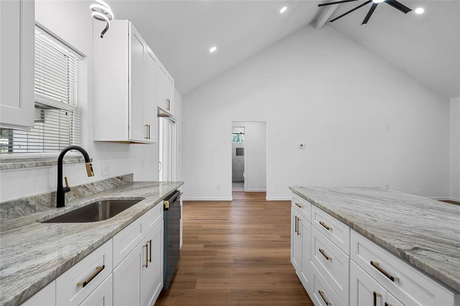Kitchen featuring white shaker cabinetry, light-toned stone countertops, a stainless steel sink with a matte black and gold-tone faucet, and wood-finish flooring Kitchen featuring white shaker cabinetry, light-toned stone countertops, a stainless steel sink with a matte black and gold-tone faucet, and wood-finish flooring
