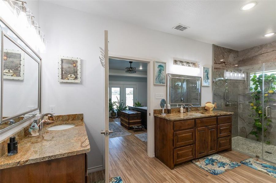 Bathroom featuring wood-finish flooring, dual vanity with granite countertops, glass-enclosed shower, and decorative stone-finish shower walls Bathroom featuring wood-finish flooring, dual vanity with granite countertops, glass-enclosed shower, and decorative stone-finish shower walls