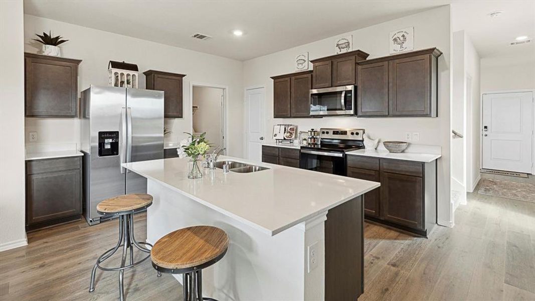 Kitchen featuring dark wood cabinetry, stainless steel appliances, light-colored countertops, and an island with an integrated sink