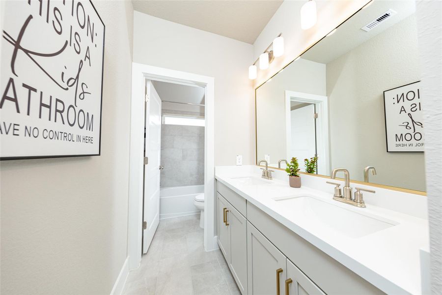 This spacious secondary bathroom features a highly functional layout highlighted by a double-sink vanity with a crisp white countertop and modern cabinetry. An expansive, wall-to-wall mirror reflects the bright overhead lighting, creating an open and airy feel. The thoughtful design includes a separate, semi-private area for the toilet and shower-tub combo, featuring contemporary gray tiling that completes this clean and efficient space.