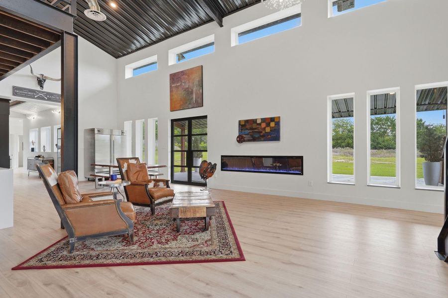 This spacious living area features high ceilings, large windows for natural light, modern decor, and a sleek electric fireplace. The open layout connects to a dining area, creating an inviting space perfect for entertaining.