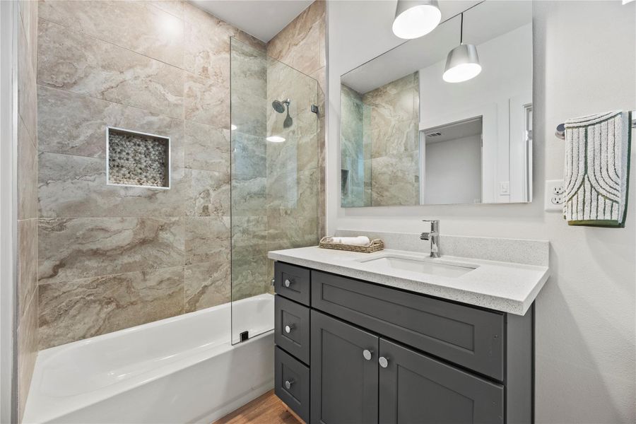 This modern bathroom boasts a sleek vanity with a quartz countertop, a large mirror, and stylish stone-tiled shower with a glass partition and built-in nook. Bright pendant lights enhance the contemporary feel.