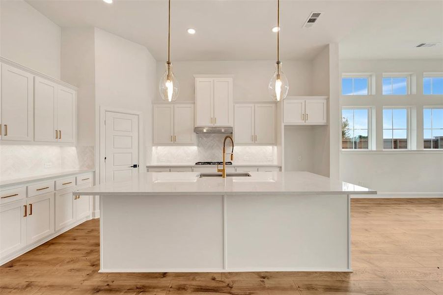 Spacious kitchen featuring a large central island with a solid surface countertop, white shaker cabinetry with gold-tone hardware, a herringbone pattern tile backsplash, and wood-finish flooring