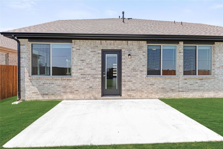 Back of single-story brick home with concrete patio, large windows, and dark glass door on green lawn