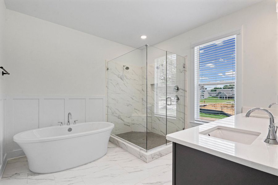 Spacious bathroom featuring a freestanding soaking tub, frameless glass shower enclosure with marble-look tile, double vanity with integrated sinks and chrome fixtures, and large window with horizontal blinds Spacious bathroom featuring a freestanding soaking tub, frameless glass shower enclosure with marble-look tile, double vanity with integrated sinks and chrome fixtures, and large window with horizontal blinds