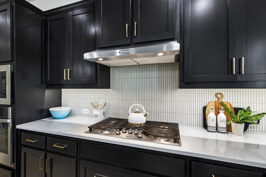Kitchen featuring black cabinetry with gold-finish hardware, light-toned countertops, and a full-height tile backsplash Kitchen featuring black cabinetry with gold-finish hardware, light-toned countertops, and a full-height tile backsplash