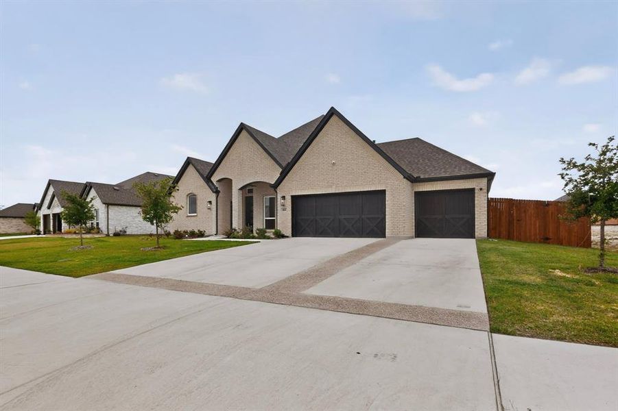 Cream brick exterior featuring dark trim, a two-car garage with X-brace doors, a covered entryway, and a concrete driveway with decorative banding Cream brick exterior featuring dark trim, a two-car garage with X-brace doors, a covered entryway, and a concrete driveway with decorative banding