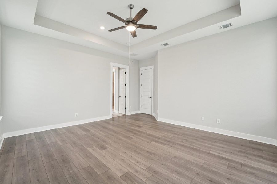 Empty room featuring a raised ceiling, wood finished floors, visible vents, a ceiling fan, and baseboards Empty room featuring a raised ceiling, wood finished floors, visible vents, a ceiling fan, and baseboards