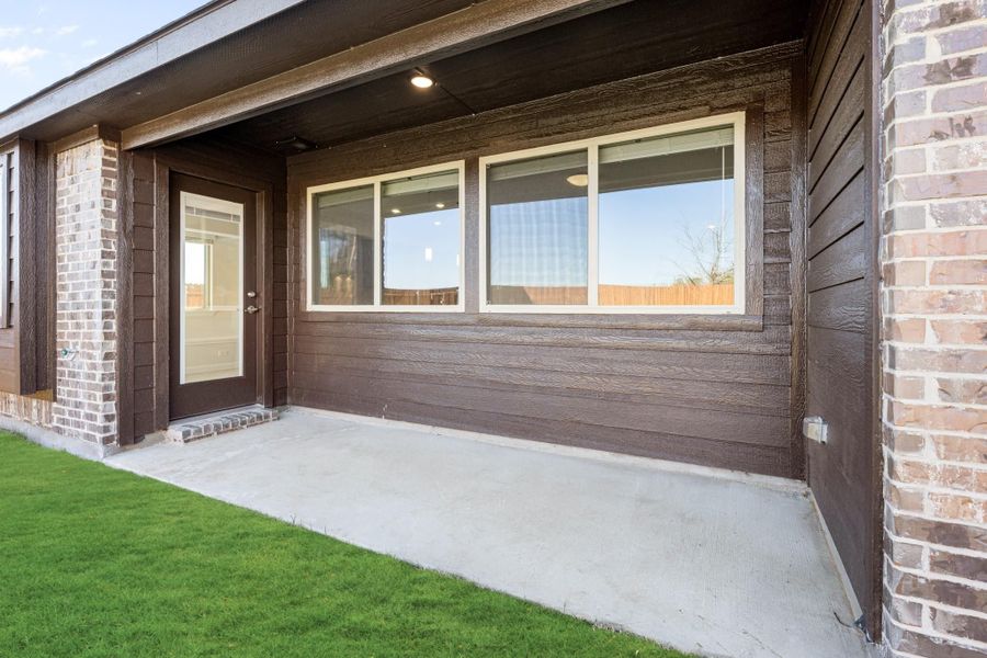 Covered back patio with concrete floor, dark wood siding, brick accents, and glass door entry beside large windows.