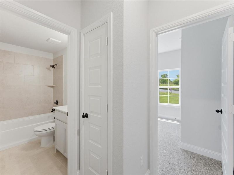 Hallway featuring a paneled door, a full bathroom with a white vanity and tub-shower combination, and a carpeted room with a window Hallway featuring a paneled door, a full bathroom with a white vanity and tub-shower combination, and a carpeted room with a window