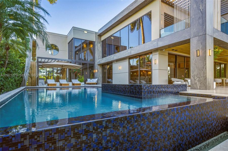 Full rear elevation view with oversized pool, blue mosaic tile spa with spillover, large-format stone deck, teak chaise lounges, stone-clad two-story facade, floor-to-ceiling glass, second-level balcony with pergola, covered loggia, wall sconces, and tropical landscaping.