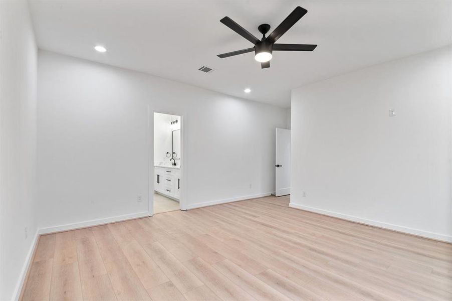 This photo shows a spacious, modern bedroom with light wood flooring, white walls, and a sleek ceiling fan. It features recessed lighting and an en-suite bathroom with a dual sink vanity.