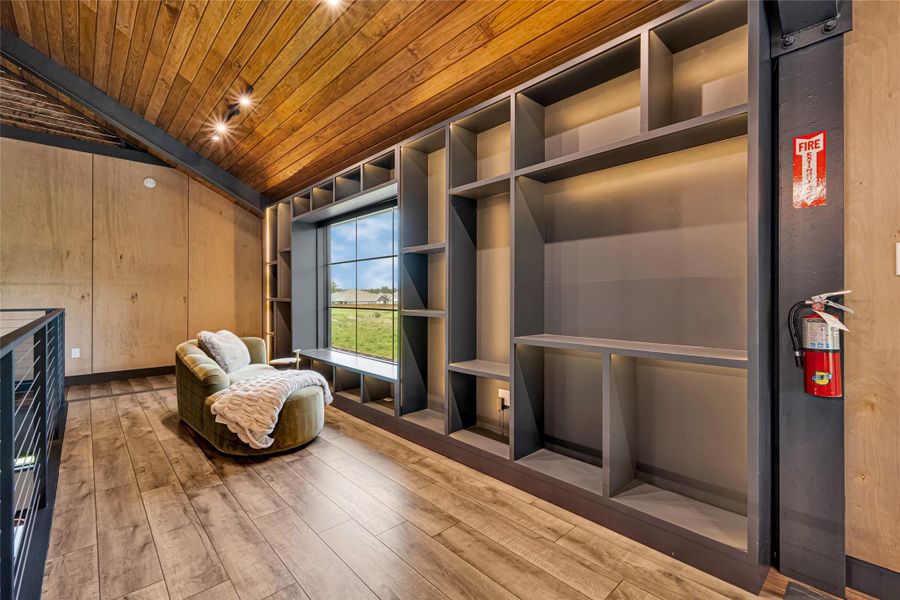 This room features a seating area with a plush chair and a blanket, ideal for relaxation. It has a modern built-in shelving unit around a large window, offering ample storage and natural light. The wood-paneled ceiling and laminate flooring add warmth and character.