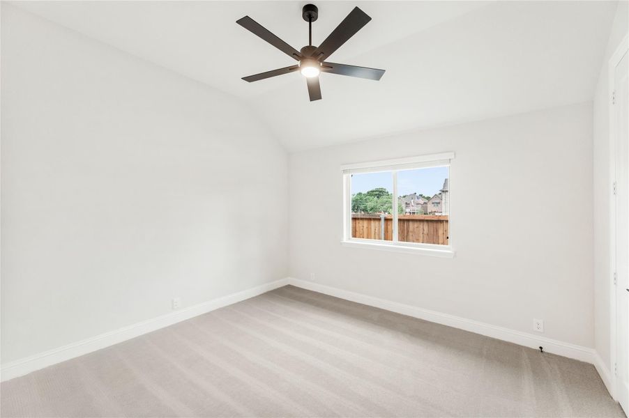 Empty bedroom with vaulted ceiling, ceiling fan, carpet flooring, and a window overlooking a backyard fence