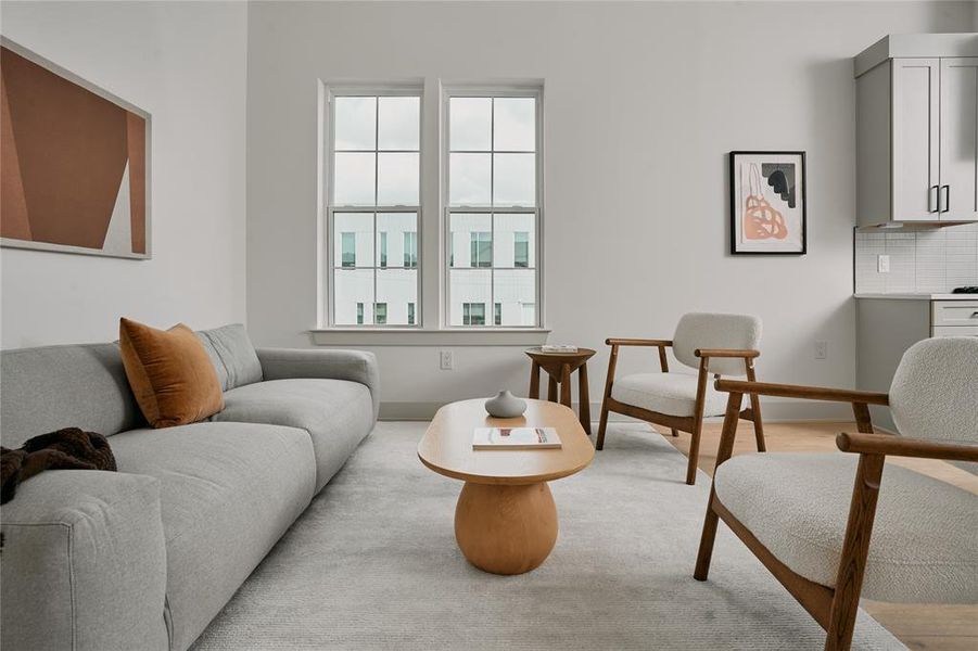 Bright living area with large windows, light wood-finish flooring, and a partial view of the kitchen featuring light cabinetry and a tiled backsplash