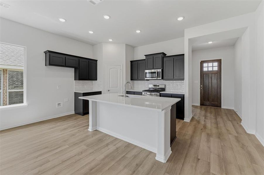 Open-concept kitchen featuring an island with a white countertop, dark cabinetry, stainless steel appliances, and wood-finish flooring Open-concept kitchen featuring an island with a white countertop, dark cabinetry, stainless steel appliances, and wood-finish flooring