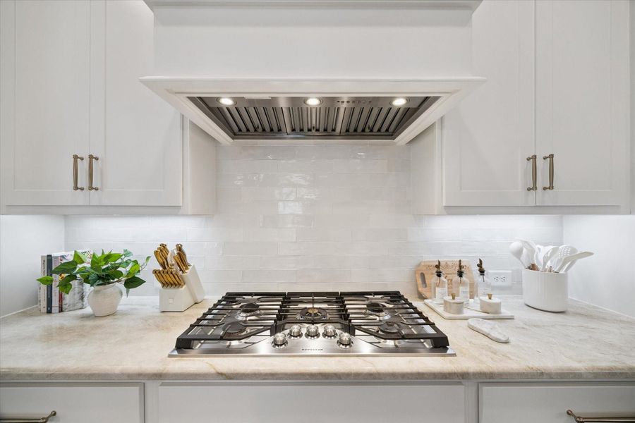 Chef’s kitchen complete with a 36" five-burner.  gas cooktop, custom vent hood, and ample counter space framed by glossy tile backsplash and quartzite countertops.